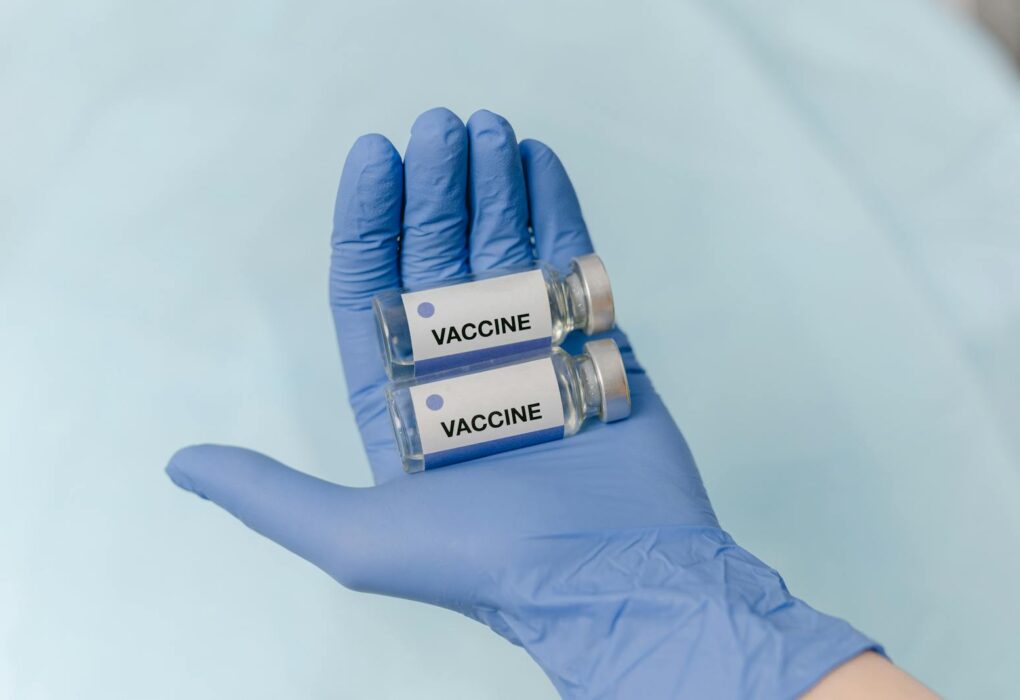 Two vaccine vials held securely in a latex-gloved hand, against a neutral background.